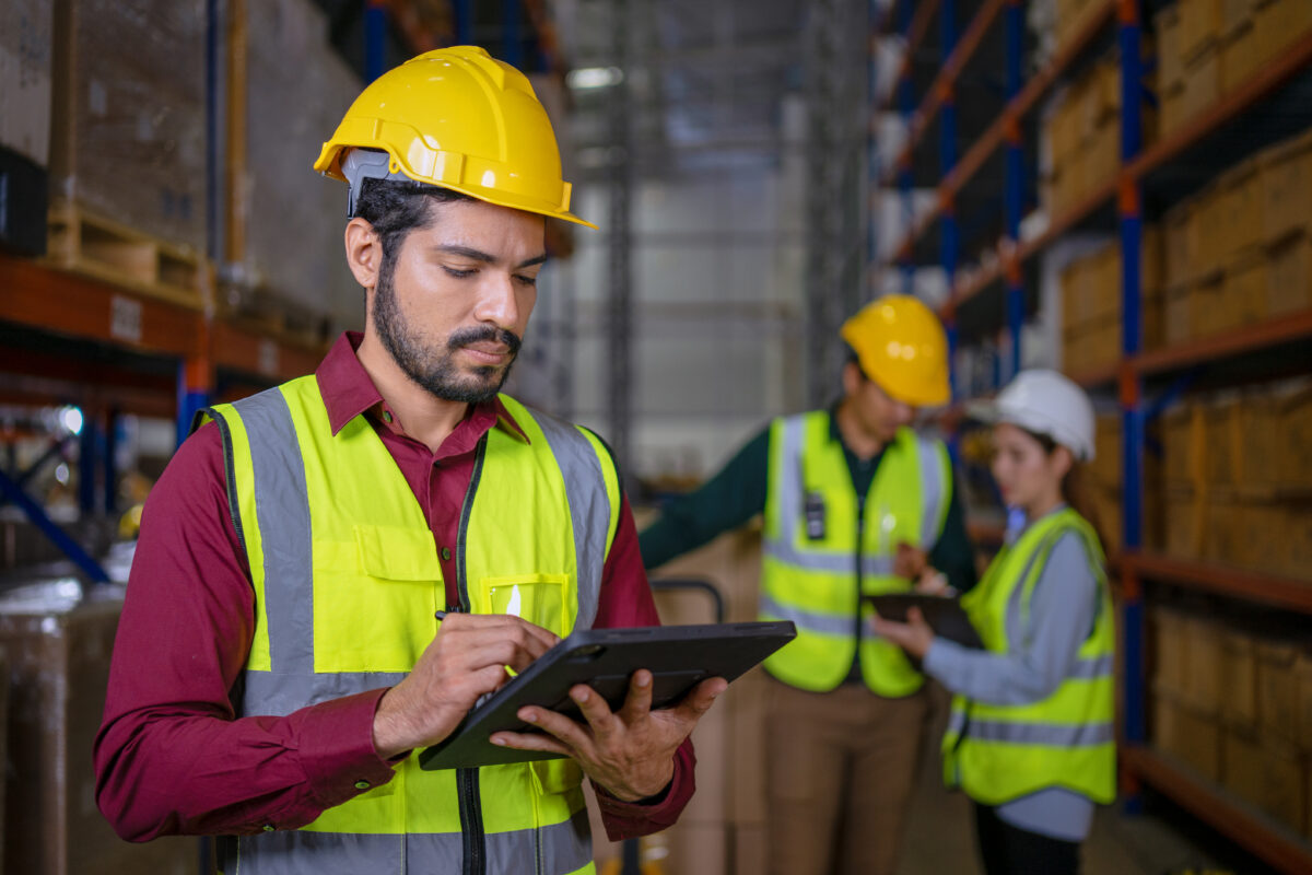 Warehouse worker in a hard hat and safety vest writing on a tablet while two colleagues work behind him