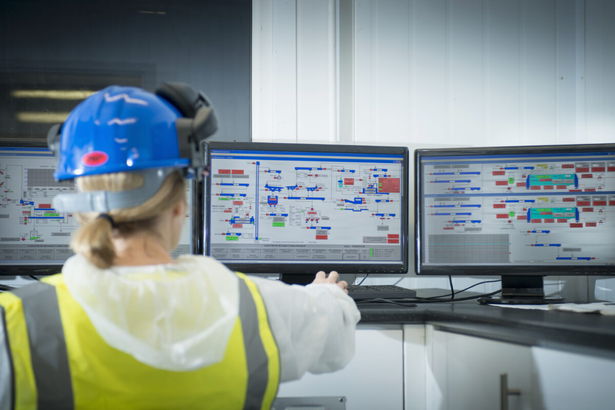 Operator in hard hat watching several monitors showing system screens in a control room