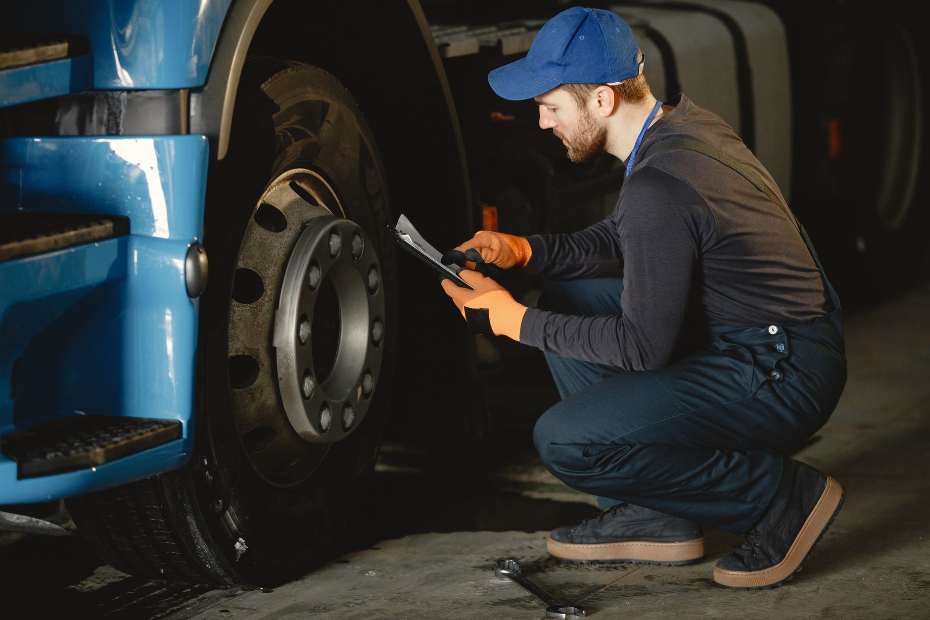 Technician in a blue cap kneeling by a truck wheel and recording an inspection on a tablet